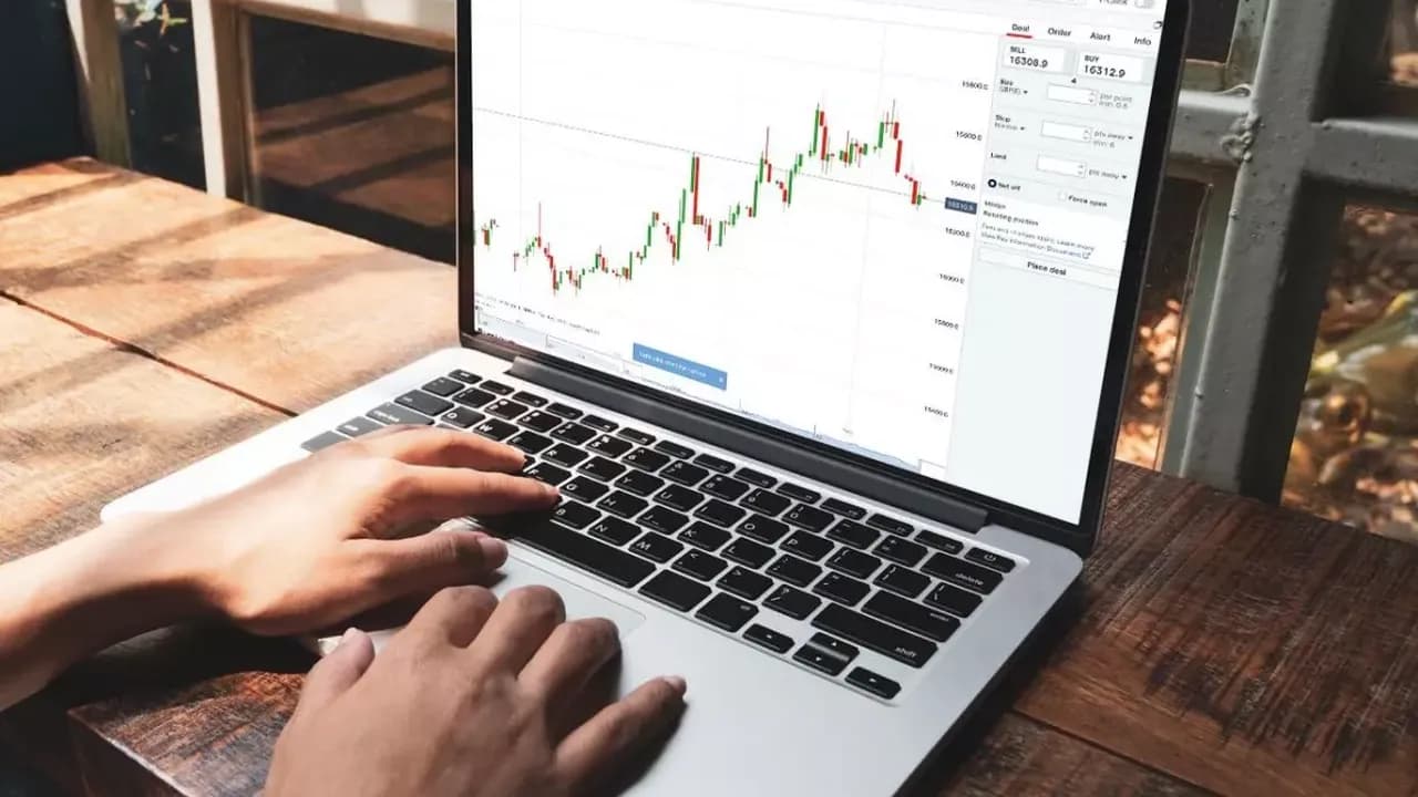 A person using a laptop displaying a financial trading chart on the screen, seated at a wooden table near a window.