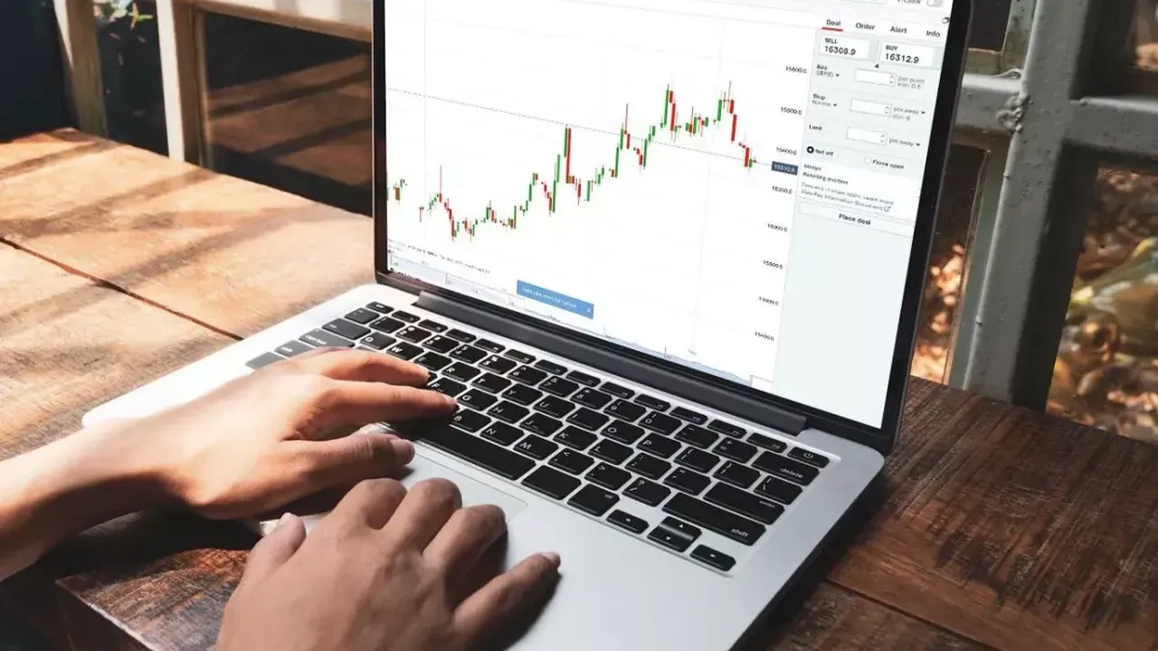 A person using a laptop displaying a financial trading chart on the screen, seated at a wooden table near a window.